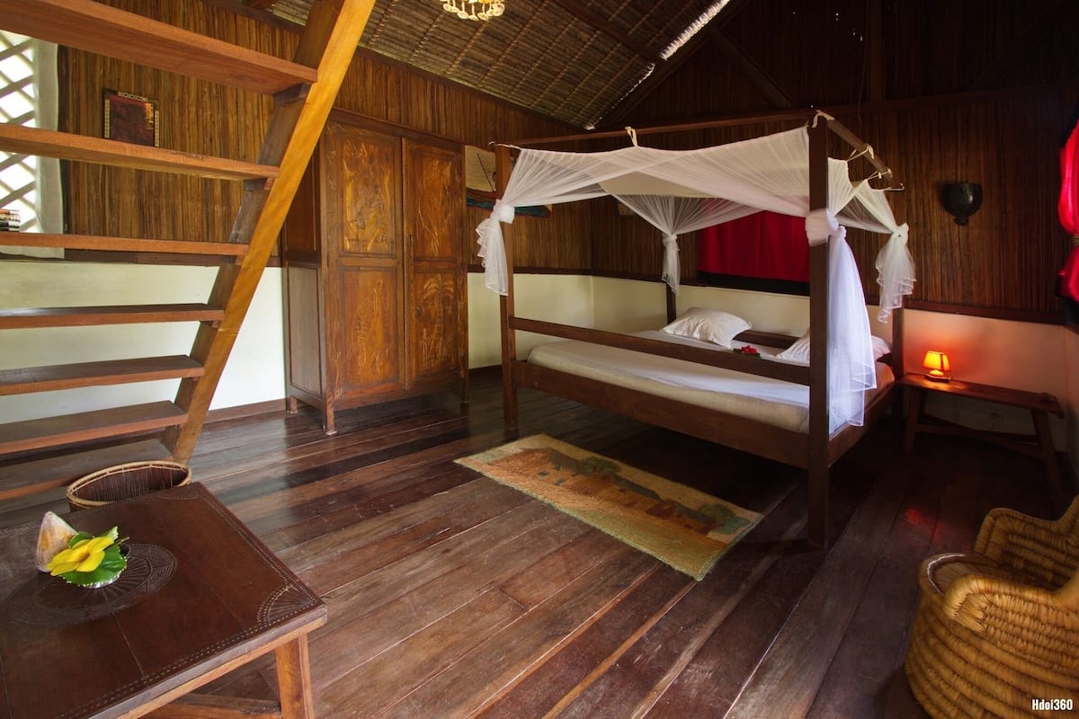 A wooden room with a bed, a small table with a flower, and a chair.