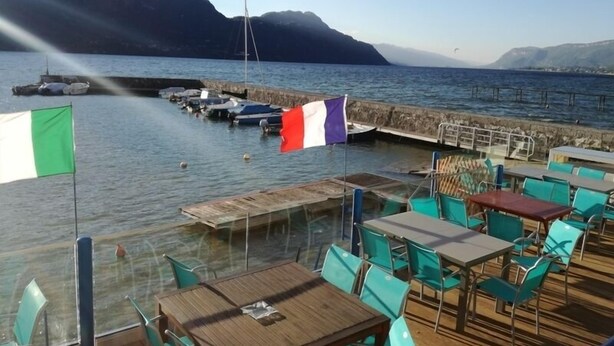 A waterfront dining area with tables and chairs, a flag, and boats docked near a pier. - Weekend Hotel Les Pieds dans l'Eau (Viviers-du-Lac)
