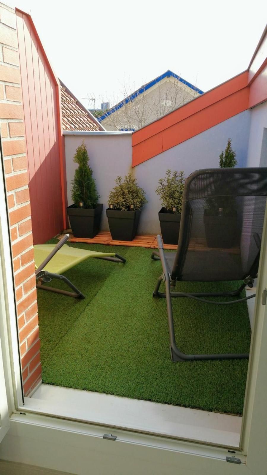Un petit espace extérieur clos, doté d’un gazon synthétique, de deux plantes en pot et d’une chaise de bureau noire.