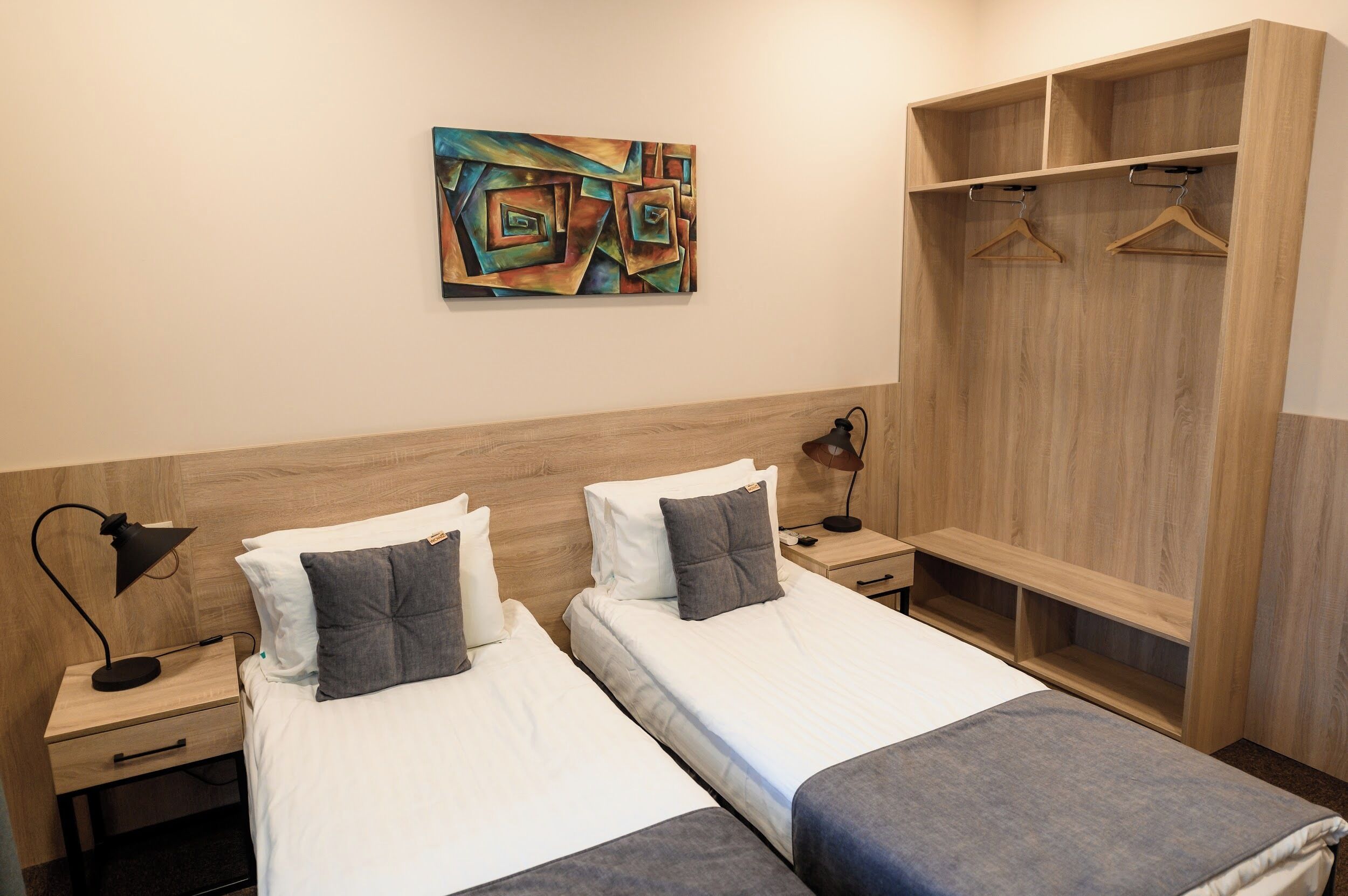 A hotel room with two beds, a wooden wardrobe, a painting on the wall, and a nightstand with a lamp.