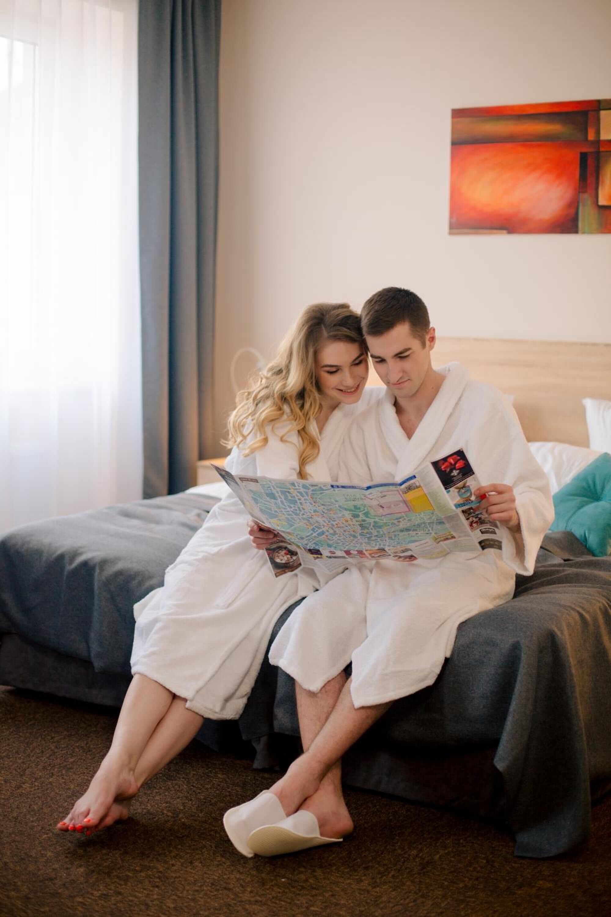 Una pareja vestida de blanco sentada en una cama con un mapa, en una habitación con un ventanal grande y un cuadro en la pared.