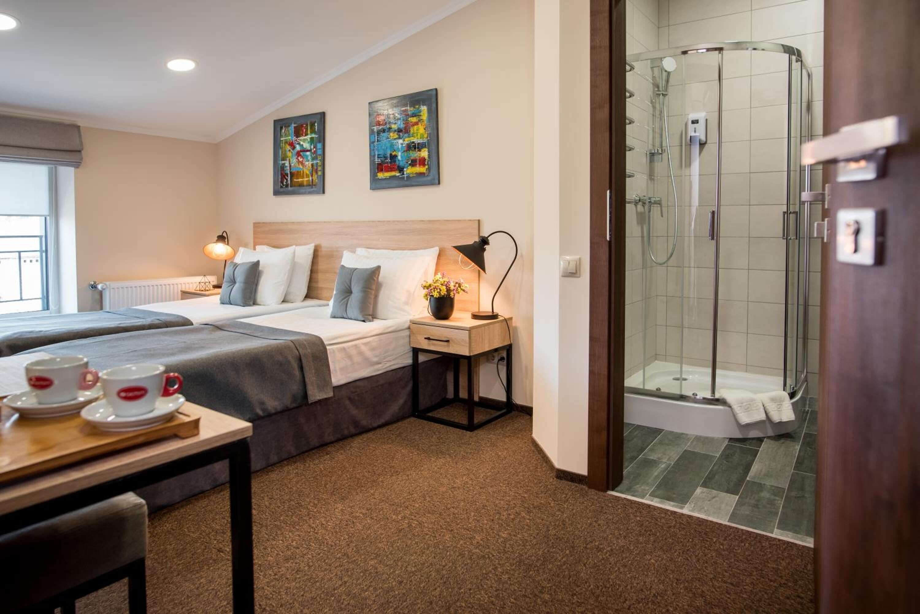 A hotel room with two beds, a small table with cups, a nightstand, a lamp, and a shower area.