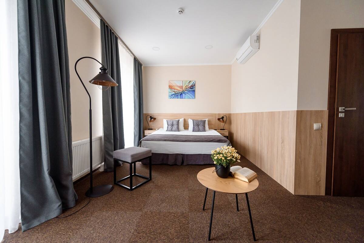 Habitación de hotel con cama, mesita de noche con un libro y flores, lámpara de pie y vistas a la ciudad.