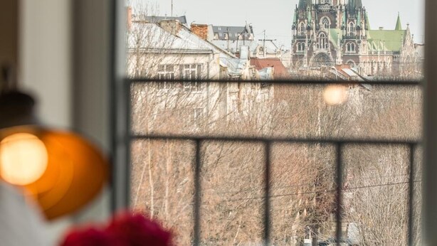 A view through a window of a building with a green roof and a tall, ornate structure with multiple spires. - Nota Bene Loft (Lviv)