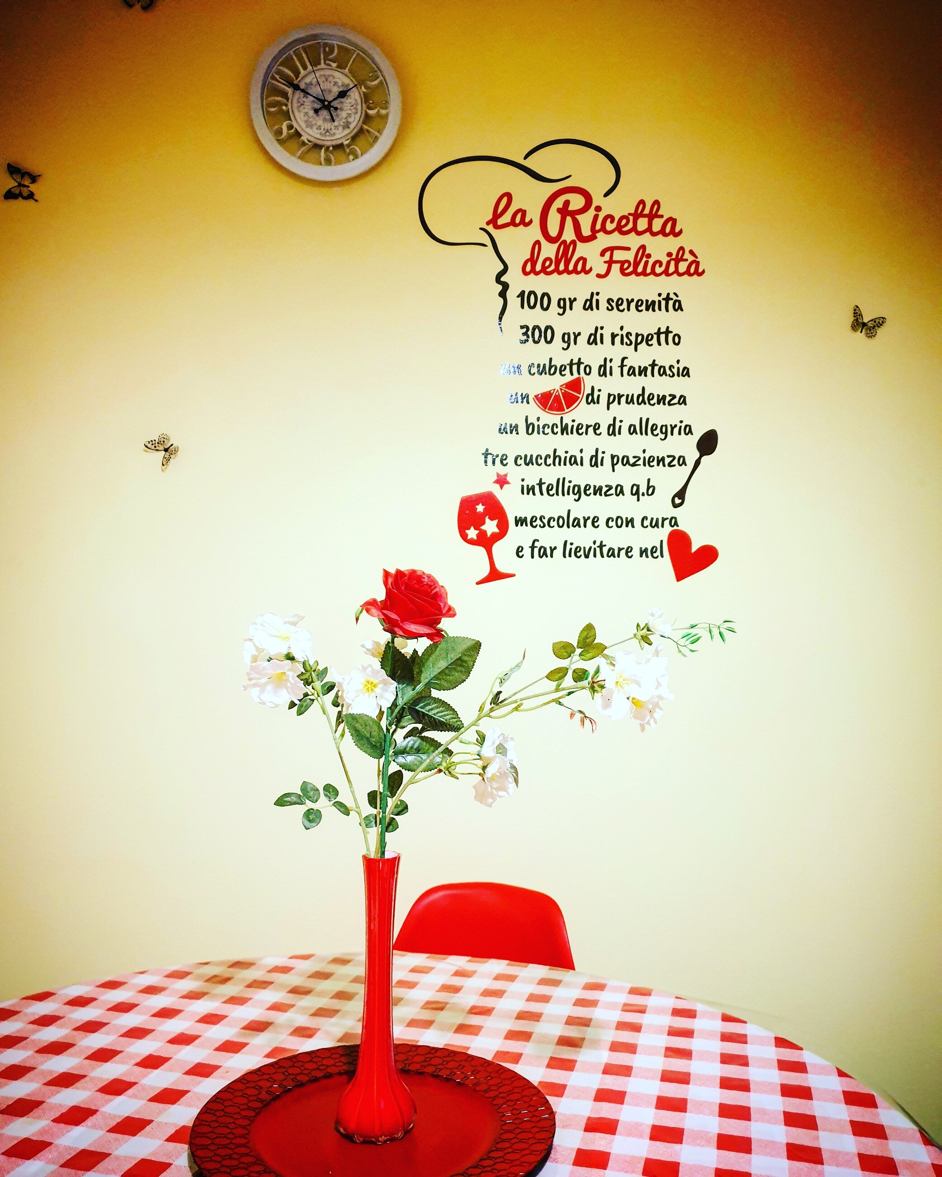 Ett bord med en röd vas med blommor, en röd stol och en väggklocka.