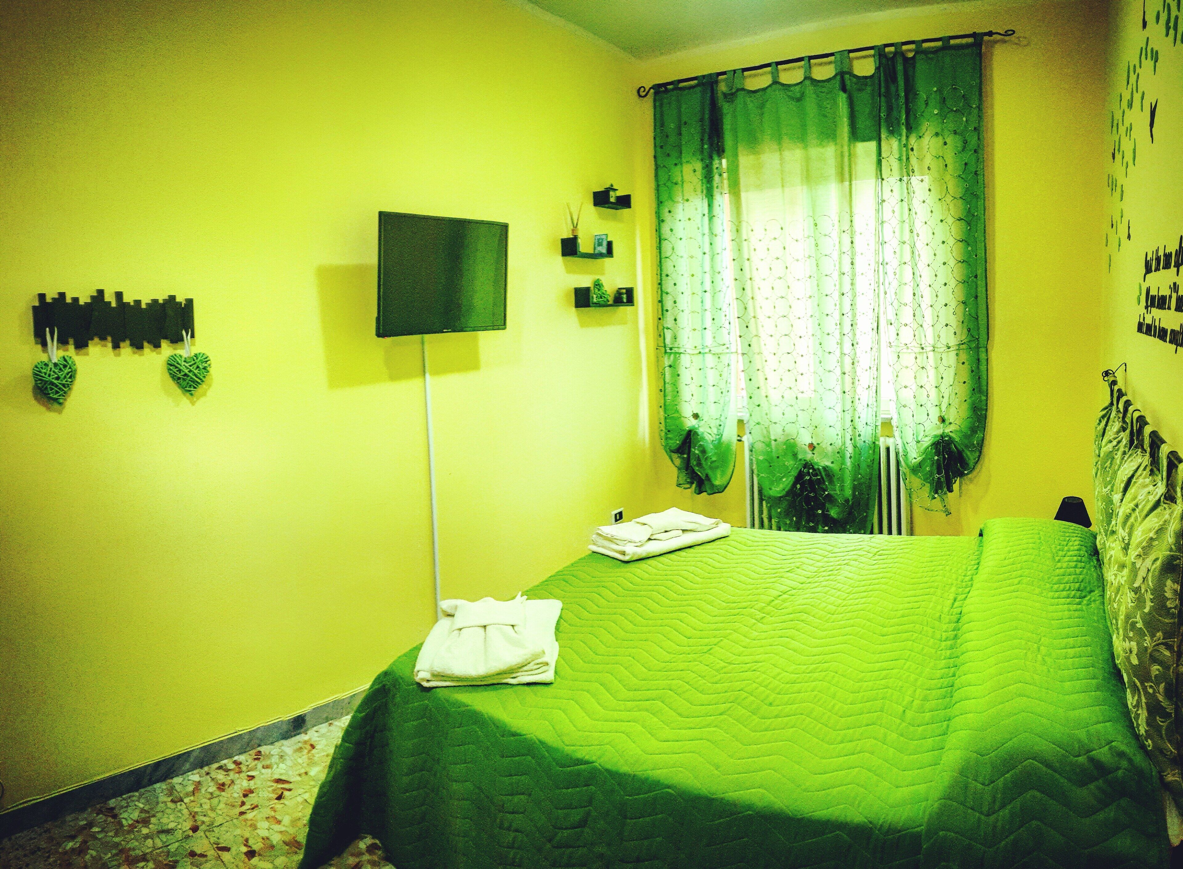 A room with a bed, a flat-screen TV mounted on the wall, and a window with a green curtain.