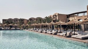 Un área junto a la piscina con sillones de descanso y sombrillas, una piscina cristalina y un edificio con varios balcones.