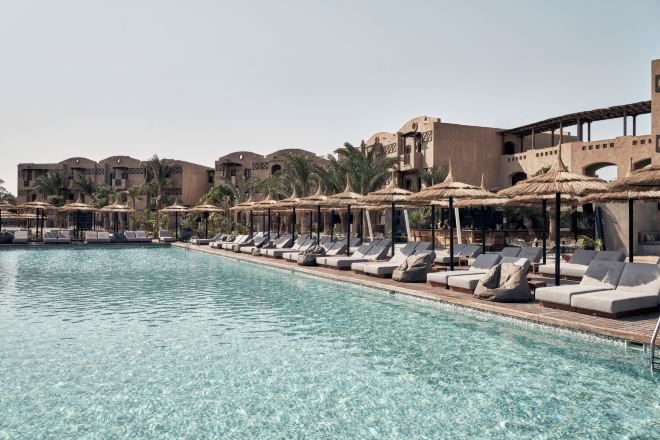 A poolside area with lounge chairs and umbrellas, surrounded by buildings with palm trees.