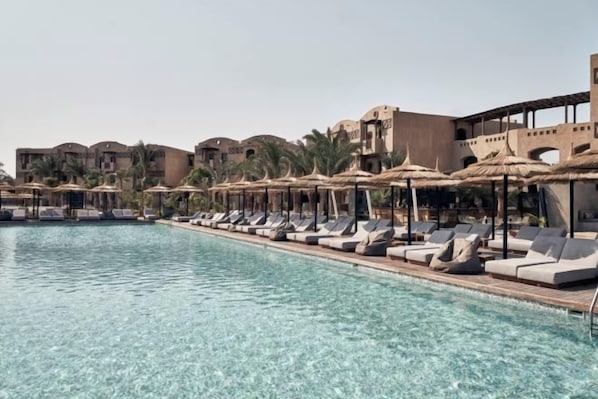 A poolside area with lounge chairs and umbrellas, surrounded by buildings with palm trees.