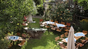 An outdoor patio area with multiple wooden tables and chairs, surrounded by lush greenery and flowering plants. - Gasthaus Sindel-Buckel Herrenhaus (Feuchtwangen)