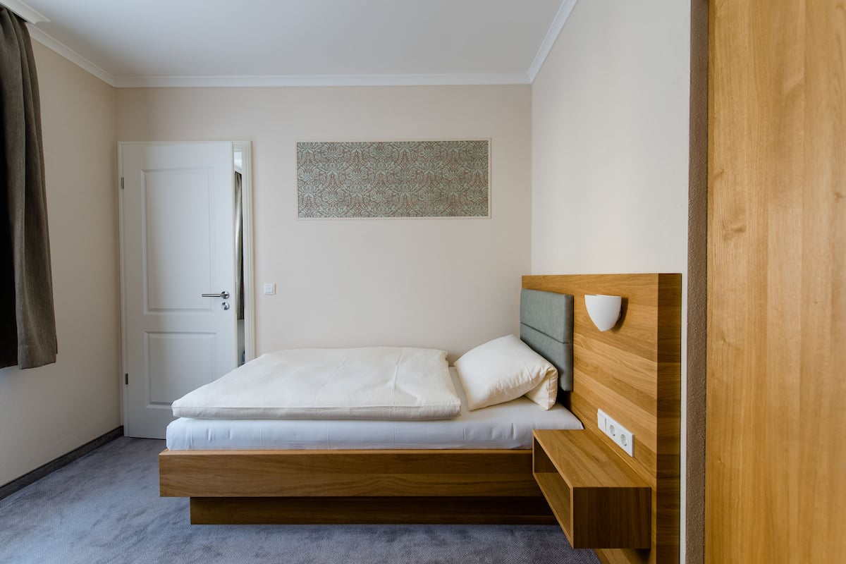 Ein Einzelbett mit Holz-Kopfstück, ein Nachttisch, eine Wandleuchte und eine Tür.