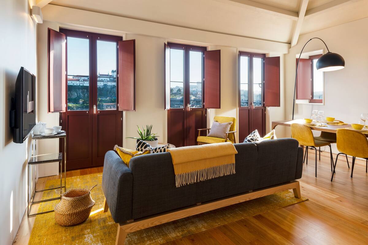 Una sala de estar moderna con un sofá, sillones amarillos, una mesa de comedor con sillas amarillas, un televisor y ventanas grandes con persianas rojas.
