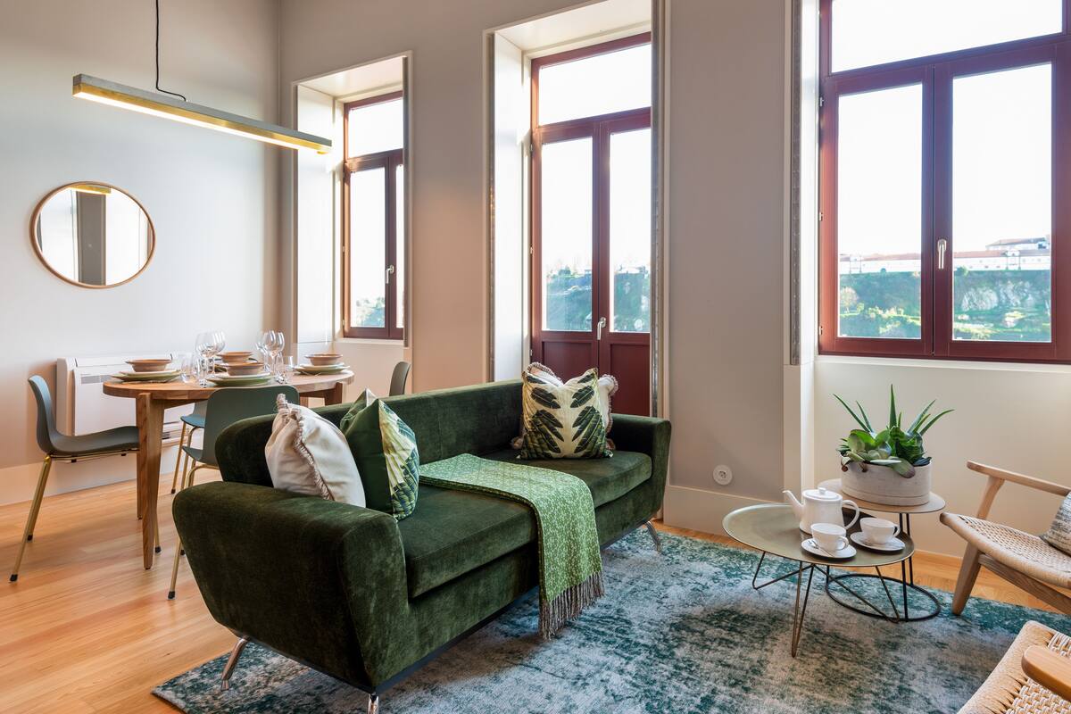Una sala de estar moderna con un sofá verde, una mesa de comedor con sillas y ventanas grandes que ofrecen vistas.