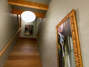 Un escalier en bois surmonté d’un luminaire moderne, un tableau encadré accroché au mur et, au fond, une œuvre d’art abstraite aux couleurs vives.