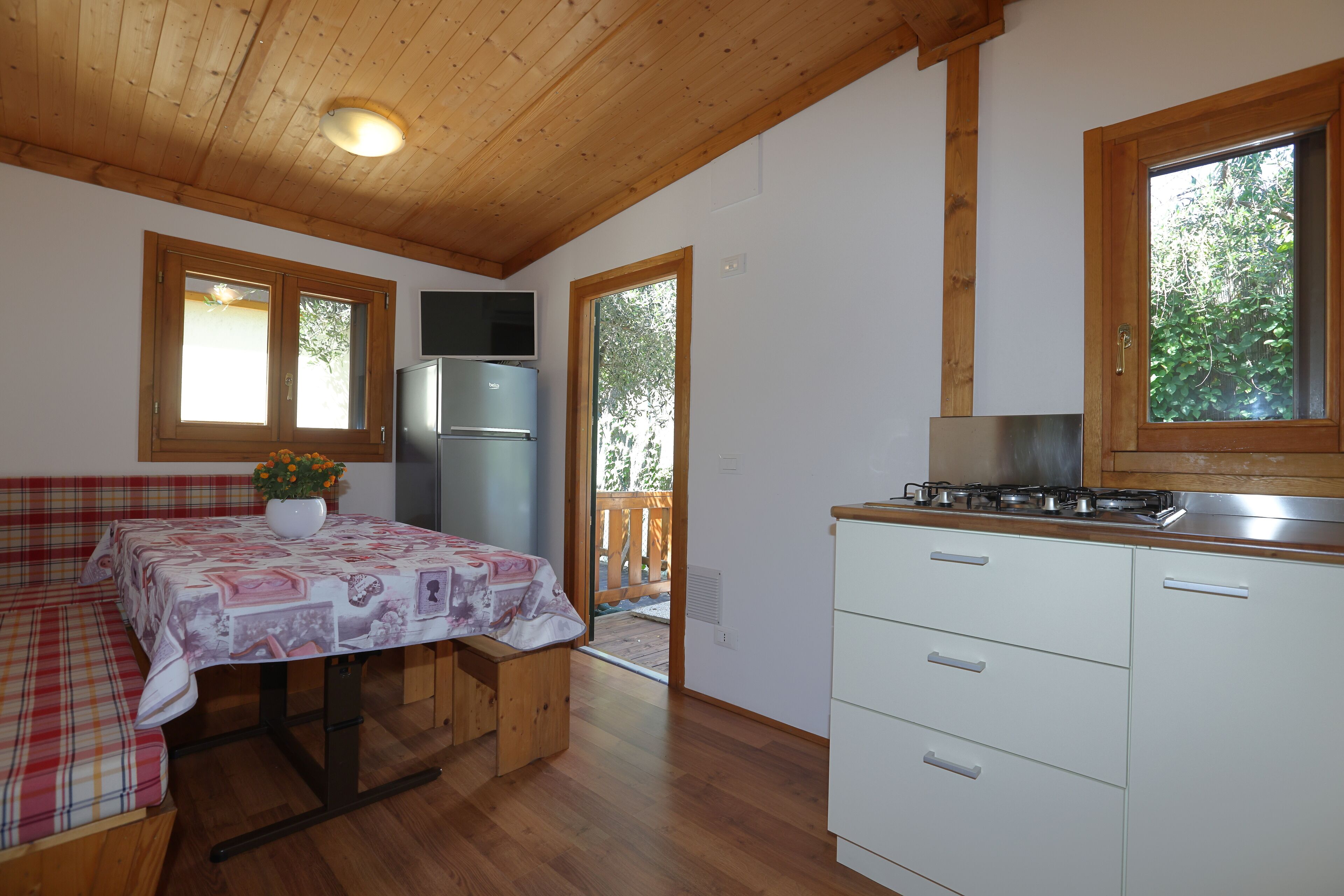 Une cuisine de taille modeste avec un plafond en bois, une table à manger recouverte d’une nappe à motifs floraux, un réfrigérateur et une fenêtre donnant sur un paysage verdoyant.