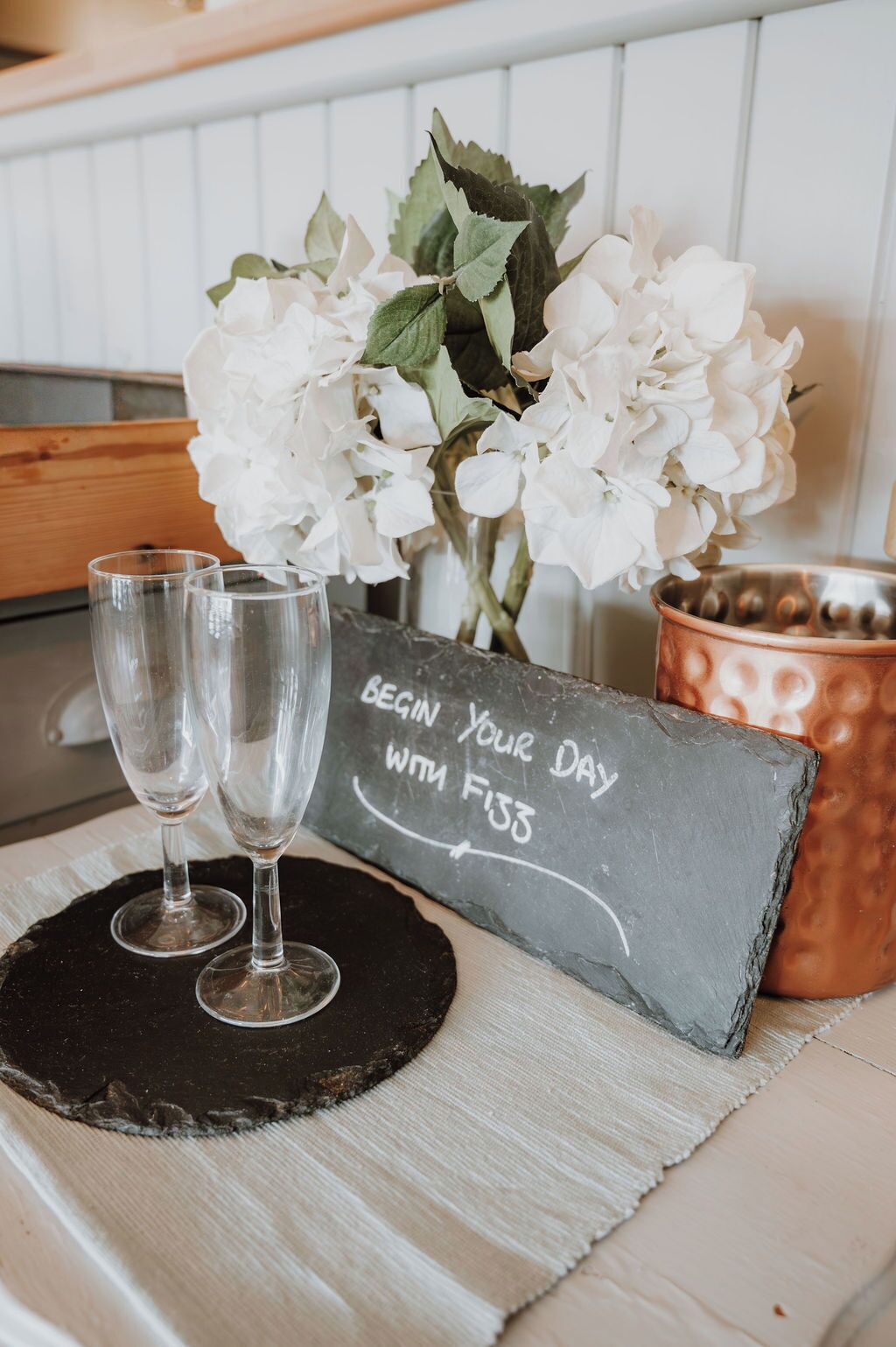 A table with two glasses, a chalkboard sign, and a vase of white flowers.