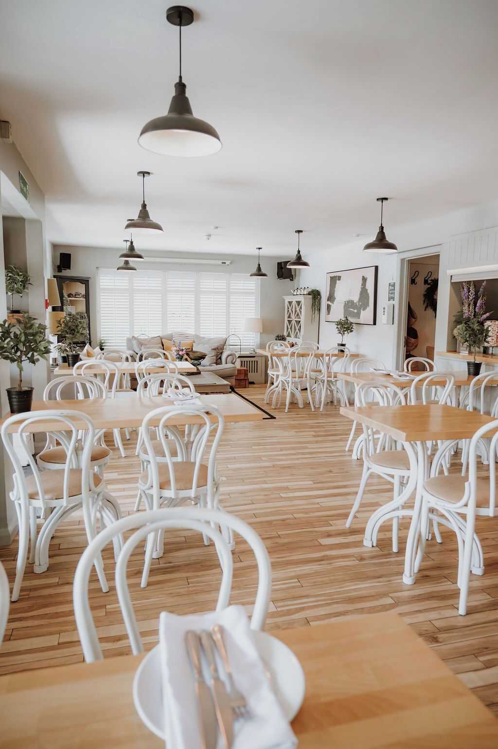 A dining area with white chairs and tables, pendant lighting, and a view of the dining room in the background.