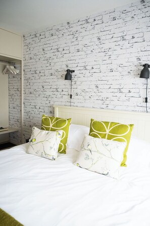 A hotel room with a white brick wall, two black wall lamps, a white bed with green and white patterned pillows, and a white nightstand with a lamp. - The Beach House (Chichester)