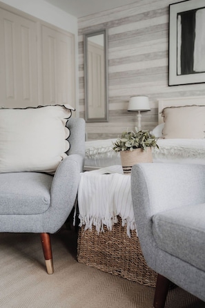 A bedroom with two grey armchairs, a white bed, a woven basket with a plant, and a mirror on the wall.