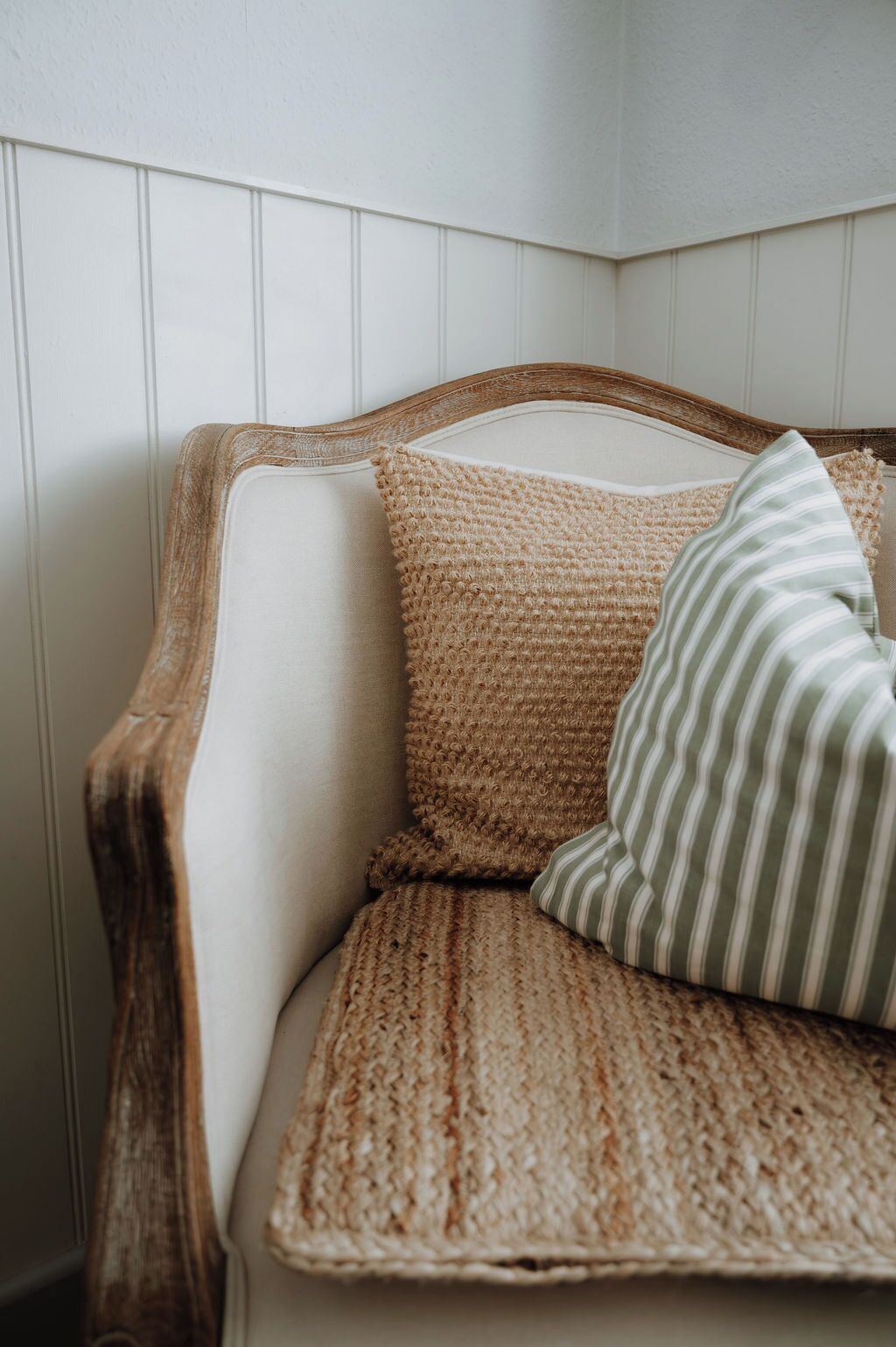 A corner of a room with a white paneled wall, a wooden-framed chair, and a woven cushion.