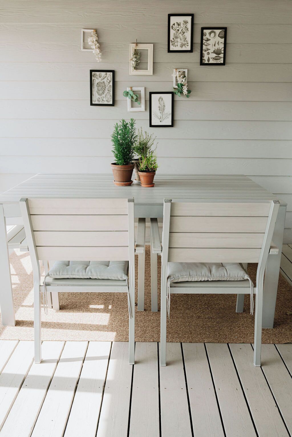 Um banco de madeira branca com duas cadeiras, uma mesa pequena e plantas em vasos num deck de madeira.