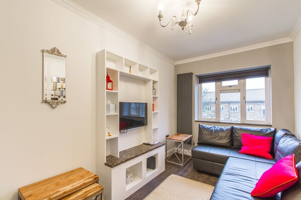 A modern living room with a flat-screen TV, a leather sofa with red cushions, a wooden coffee table, and a chandelier.