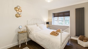 A bedroom with a white bed, a nightstand with a mirror, a lamp, and a window with blinds. - Luxury London Apartment 5-double rooms (London)
