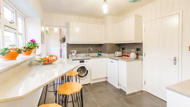 A modern kitchen with white cabinets, a washing machine, and a countertop with stools. - Luxury London Apartment 5-double rooms (London)