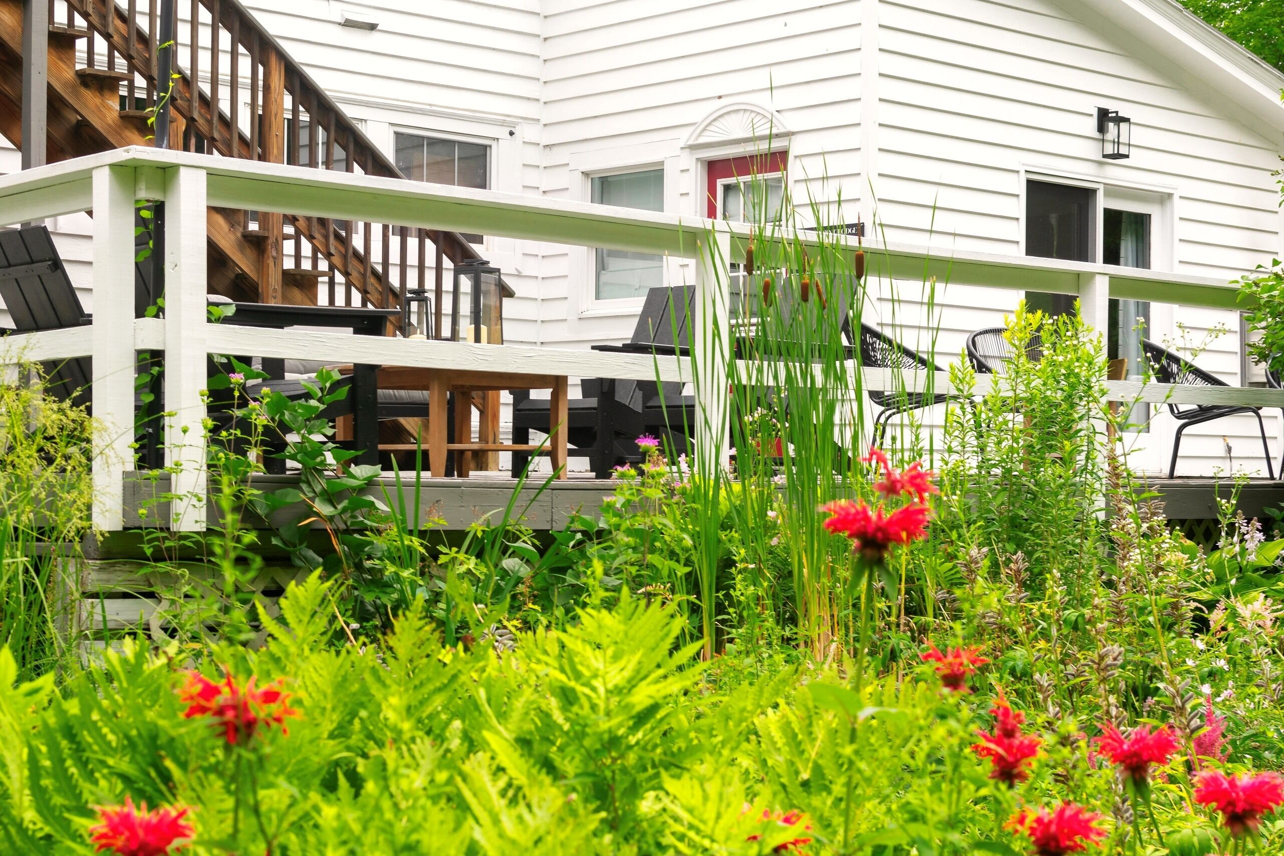 A white house with a wooden deck, outdoor seating, and a garden with red flowers.