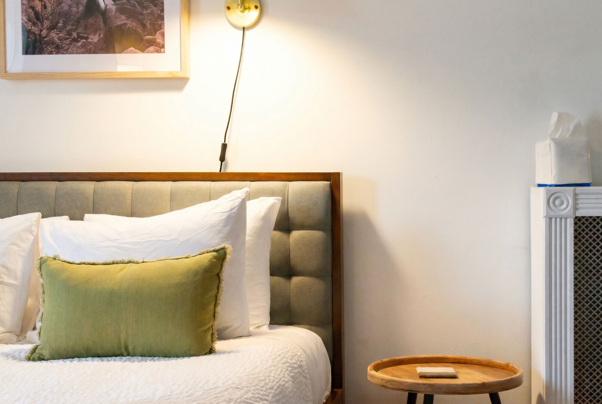 Una cama bien hecha con cabecera, una mesita de noche con un libro y una lámpara de pared.