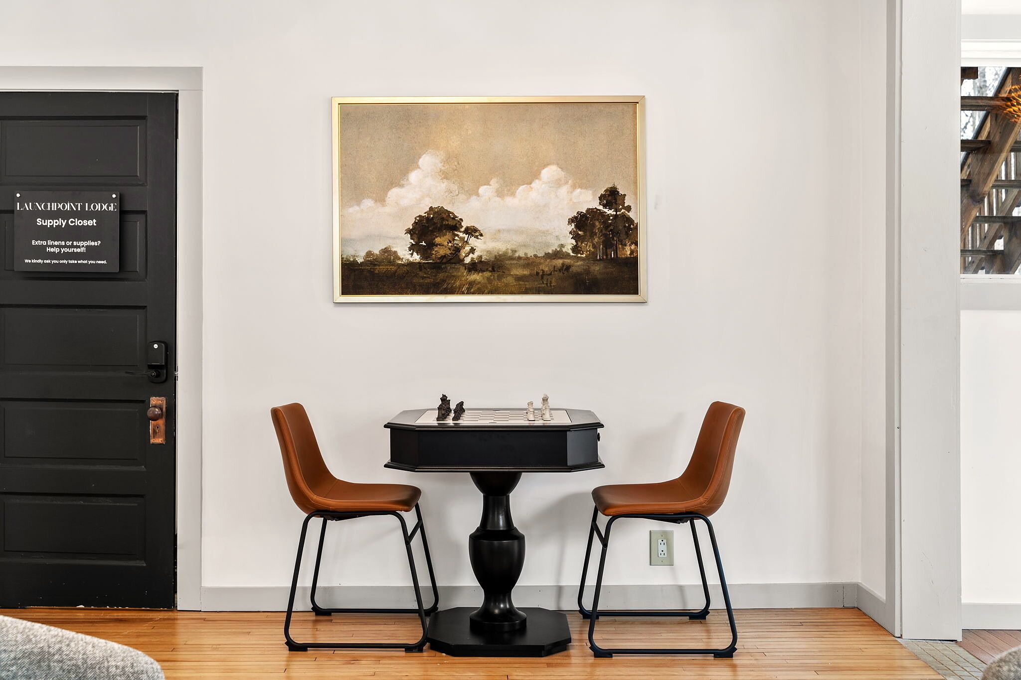 A room with a black door labeled 'LAUNCHPOINT LODGE Supply Closet,' a framed landscape painting, a chessboard, and two brown chairs.