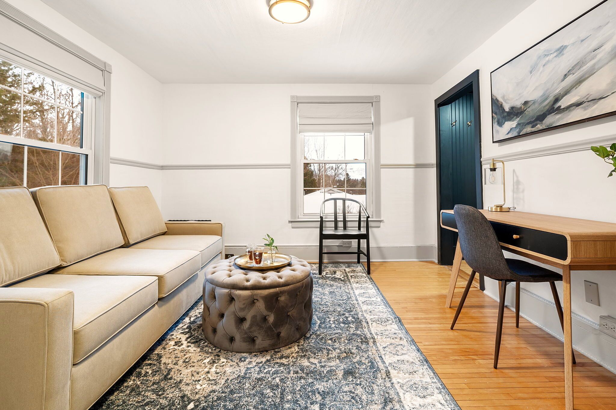 A modern living room with a beige sofa, a round ottoman, a wooden desk, and a chair.