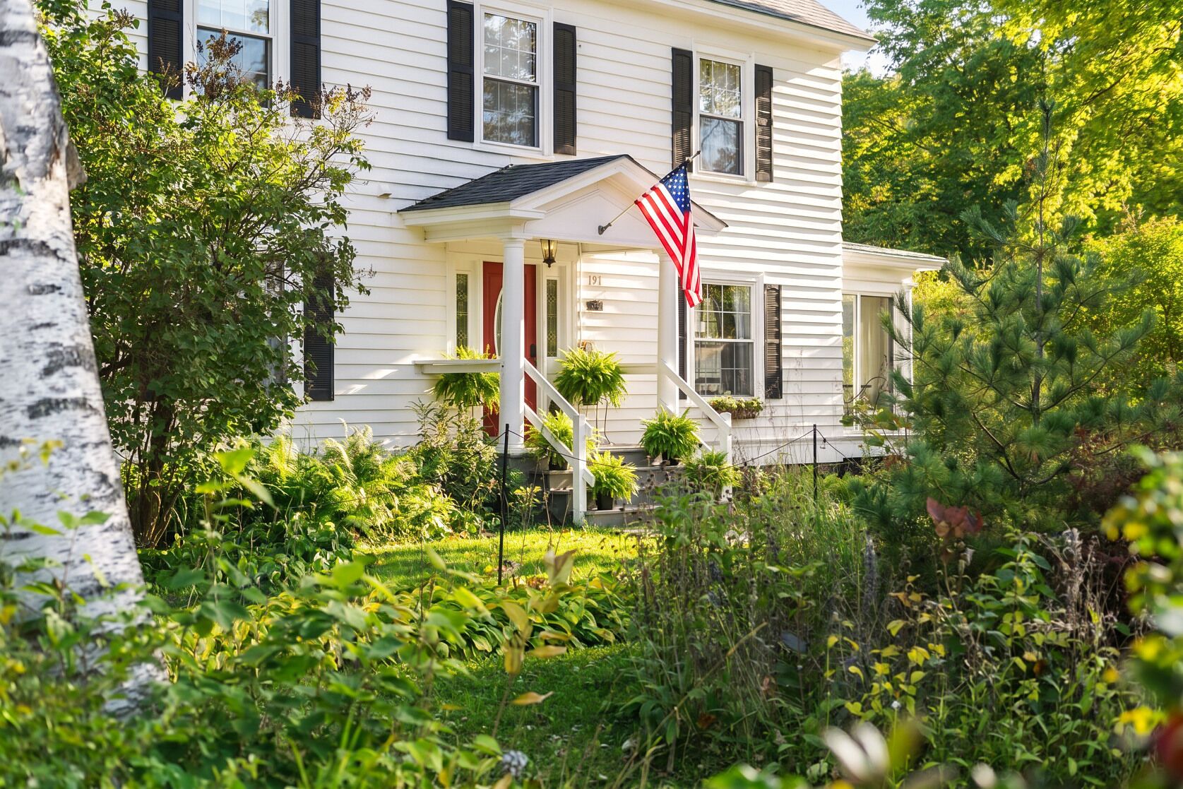 A white house with black shutters, a front porch, and an American flag.