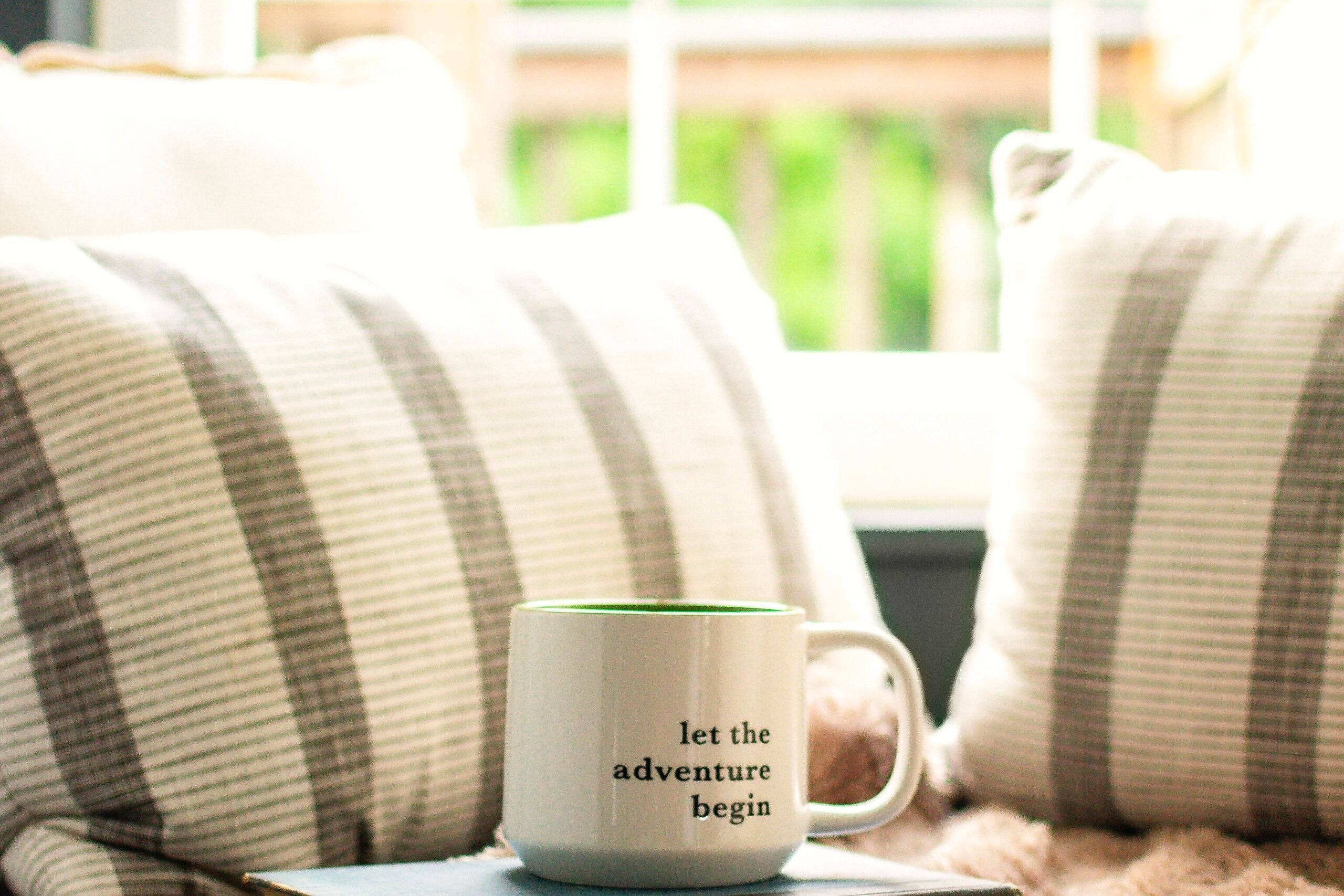A white mug with black text on a coaster, placed on a couch with striped pillows.