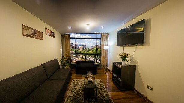 A living room with a sofa, a TV, and a view of a cityscape through large windows. - Yuyay Apartaments Cusco (Cusco)