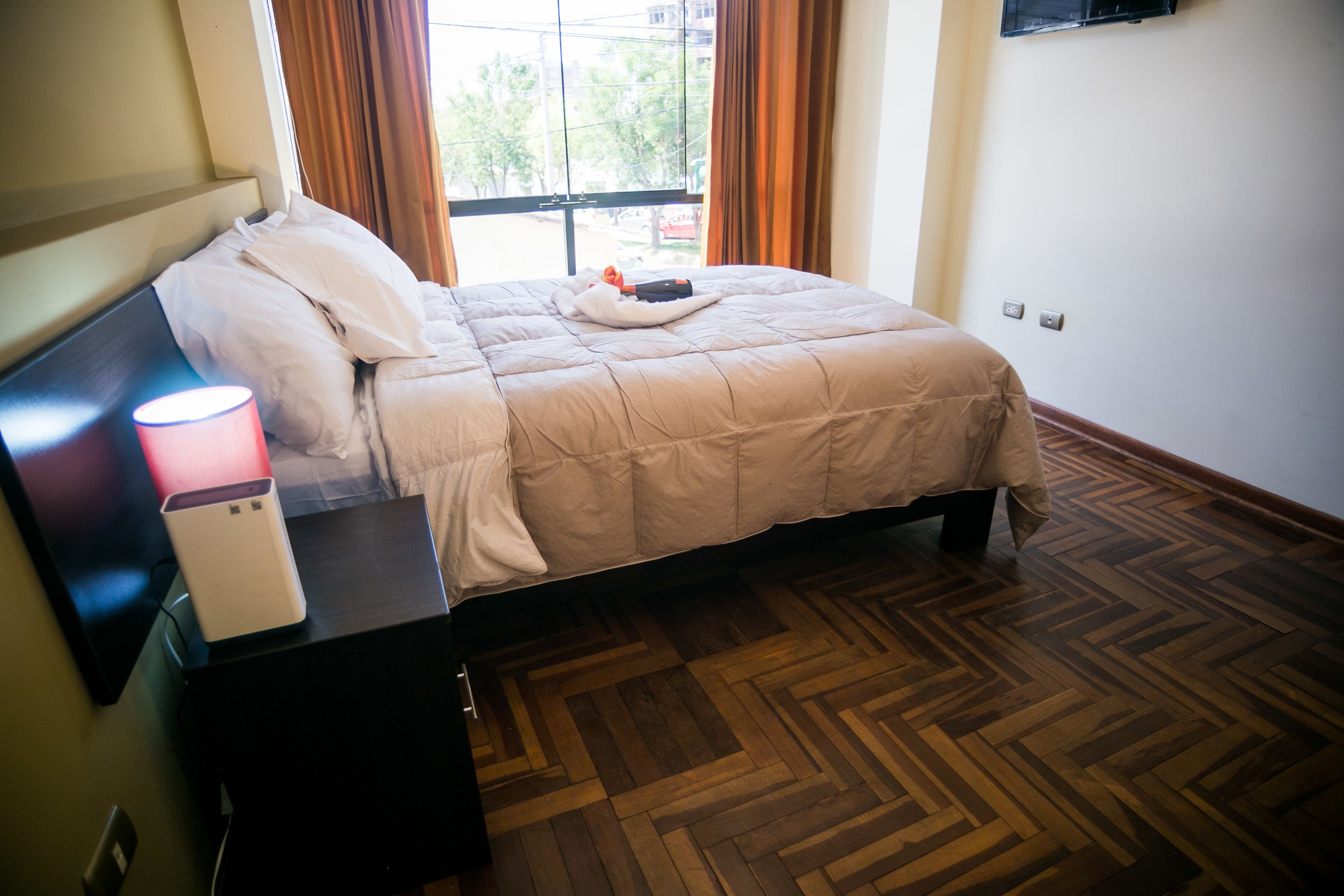 A neatly made bed with a white comforter and pillows, a bedside table with a lamp and a white device, and a wooden parquet floor.