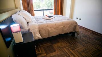 A neatly made bed with a white comforter and pillows, a bedside table with a lamp and a white device, and a wooden parquet floor.