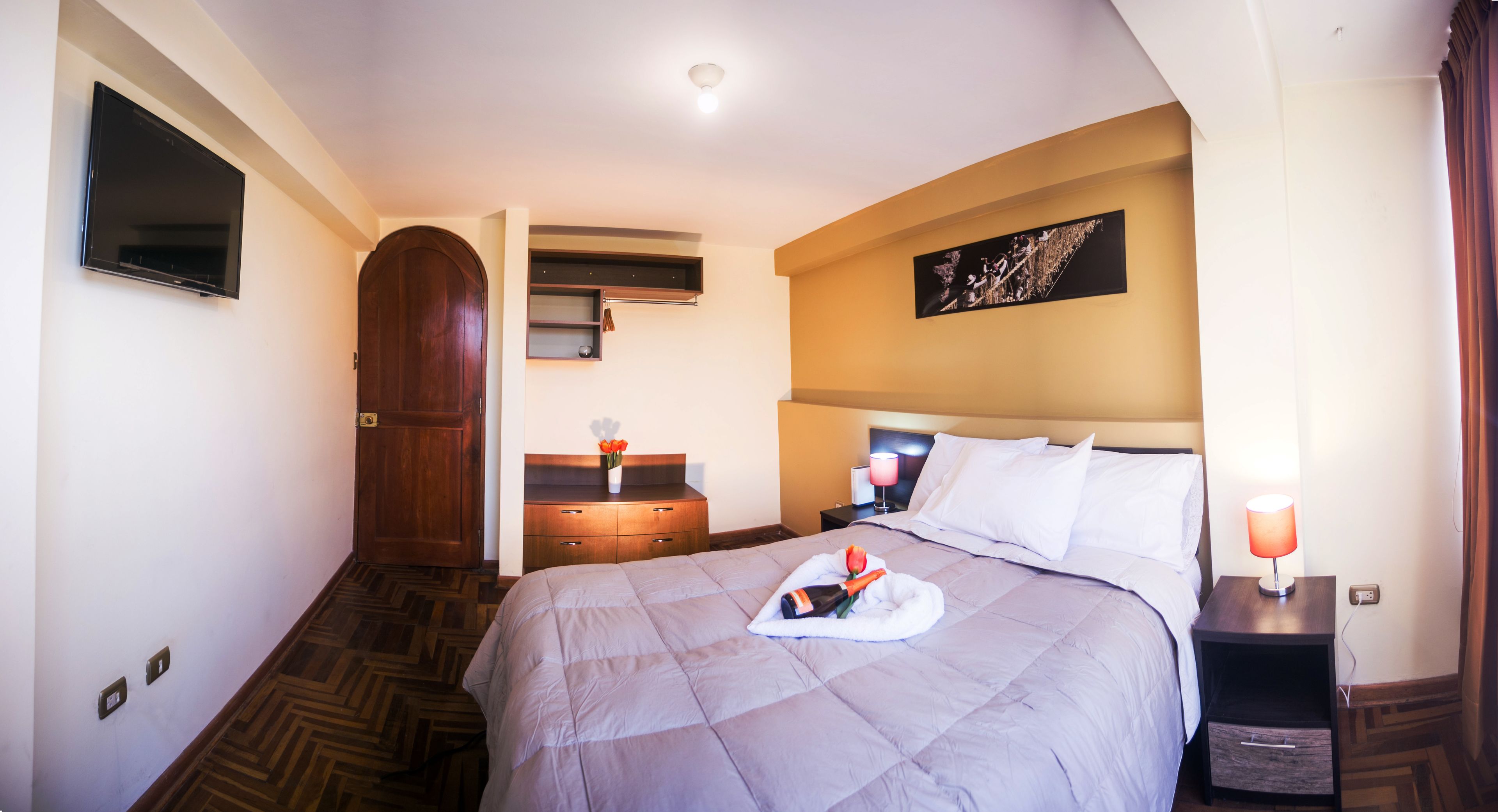 A hotel room with a bed, a nightstand, a TV mounted on the wall, a wooden door, and a painting on the wall.
