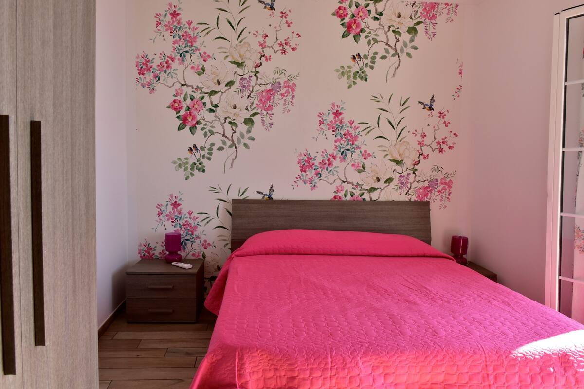 A bedroom with a wooden wardrobe, a bed with a pink quilt, and floral wallpaper.