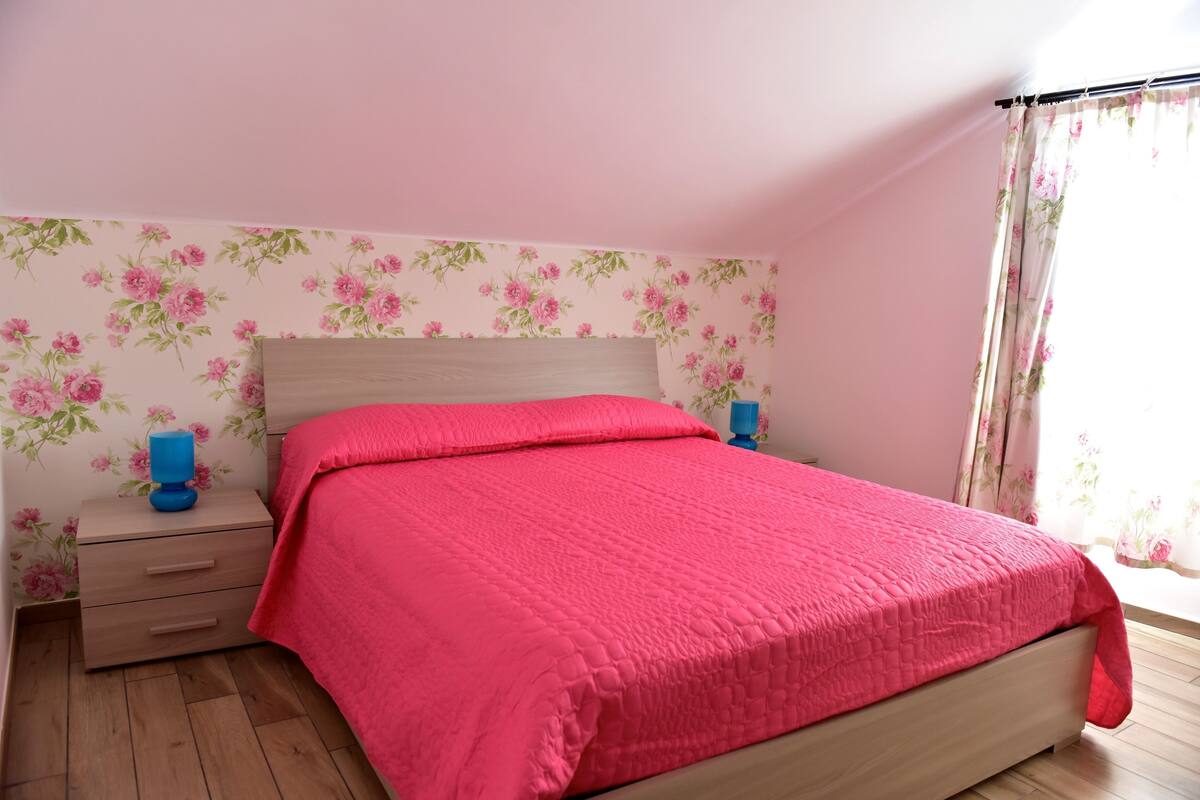 A bedroom with a wooden bed, a pink quilt, floral wallpaper, and a nightstand with a blue lamp.