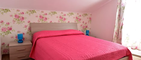 Quarto com cama de madeira, colcha rosa, papel de parede floral e mesa de cabeceira com abajur azul.