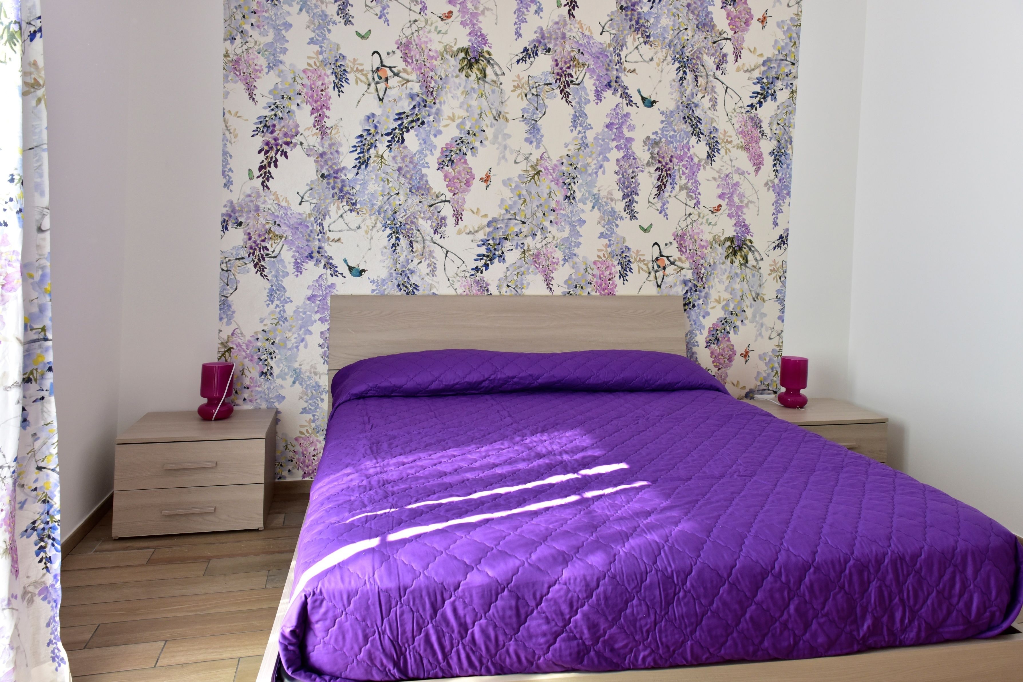 Un dormitorio con cama de madera, ropa de cama morada, papel tapiz floral y dos mesitas de noche con velas rojas.
