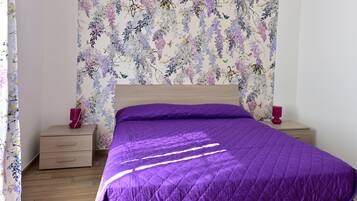 Un dormitorio con cama de madera, ropa de cama morada, papel tapiz floral y dos mesitas de noche con velas rojas.
