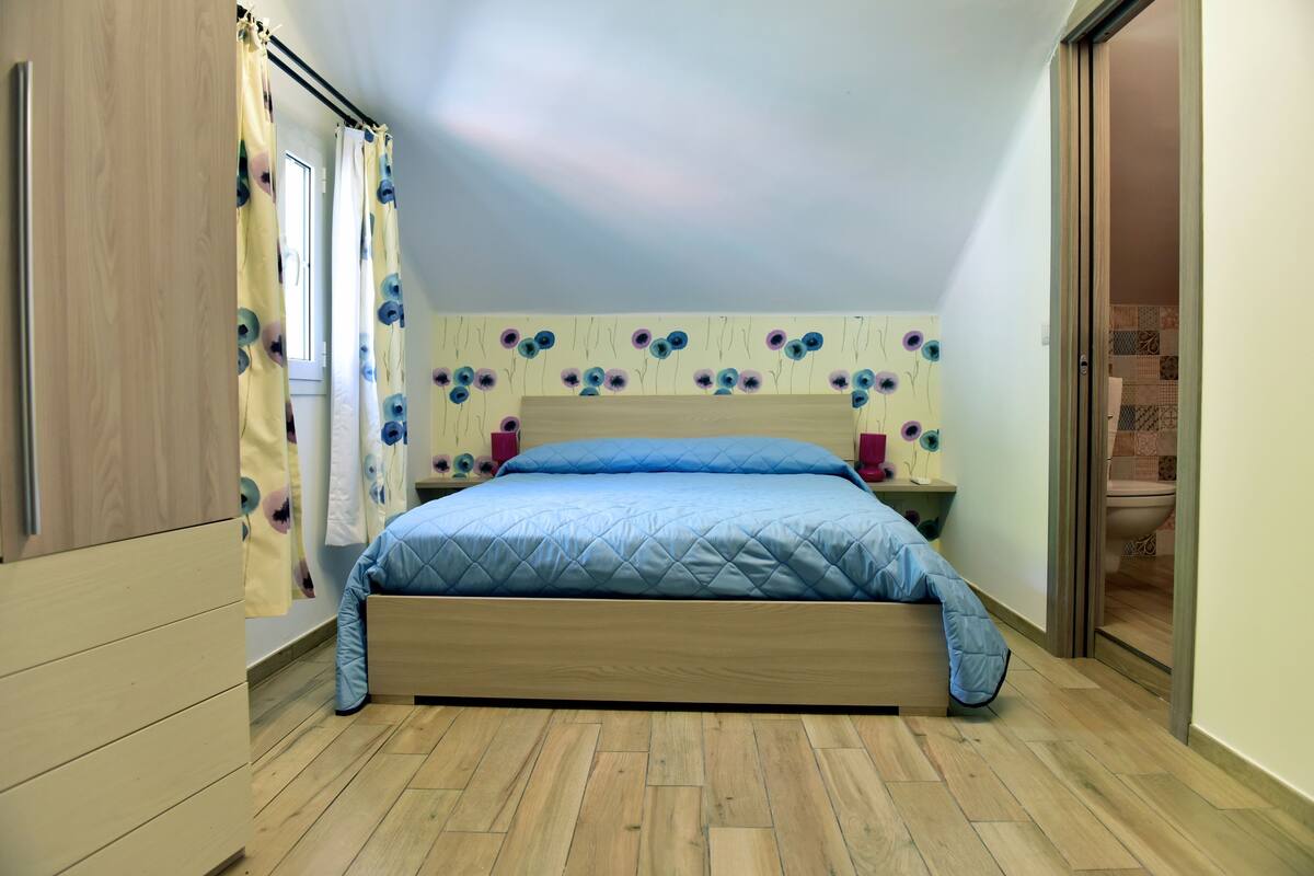 A bedroom with a wooden bed, a nightstand, a window with curtains, and a wall with a floral pattern.