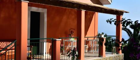 A building with a stone base and orange walls, featuring a covered porch with a metal railing and a staircase.