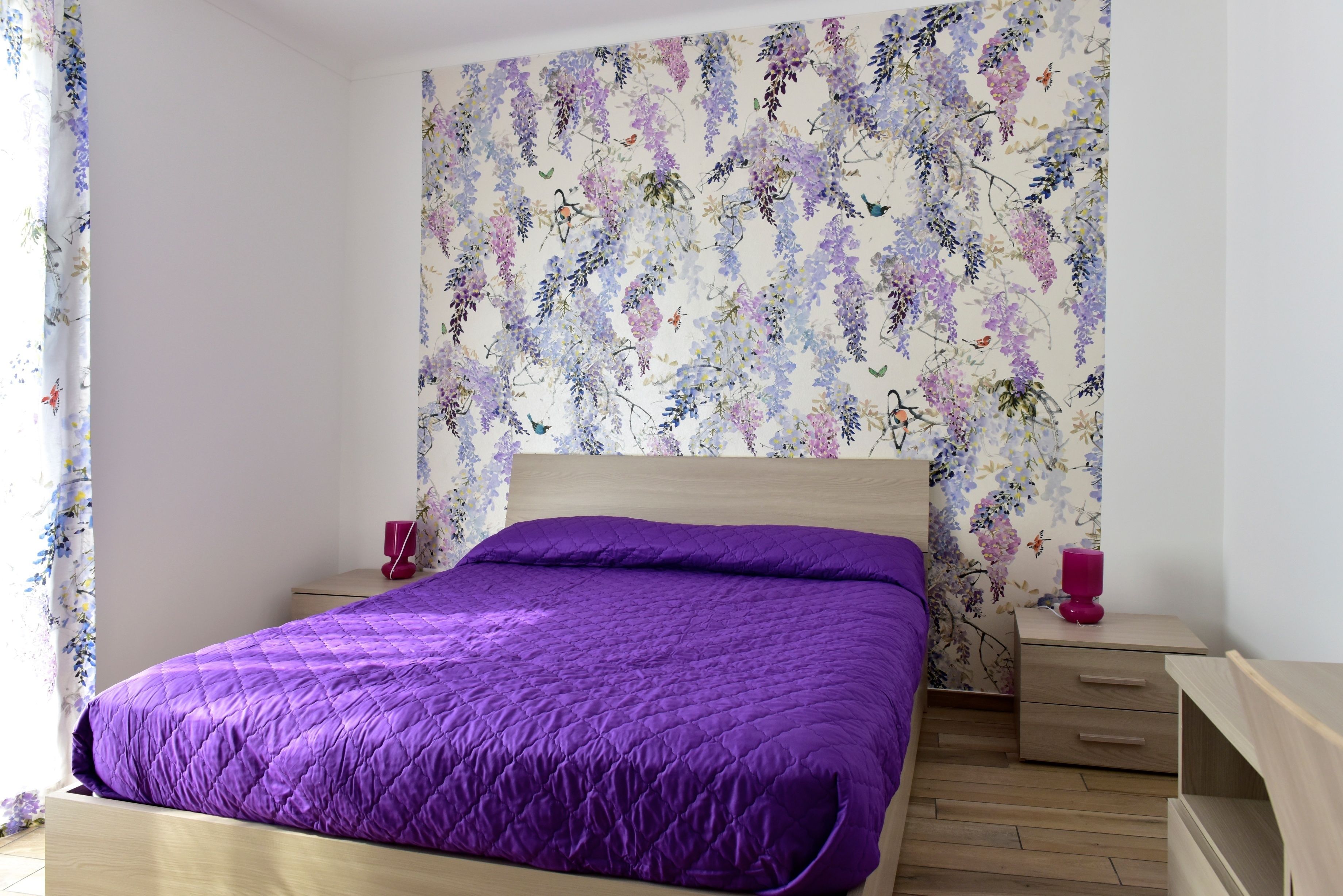 A bedroom with a purple bedspread, floral wallpaper, a wooden headboard, a nightstand with a pink lamp, and a wooden dresser.