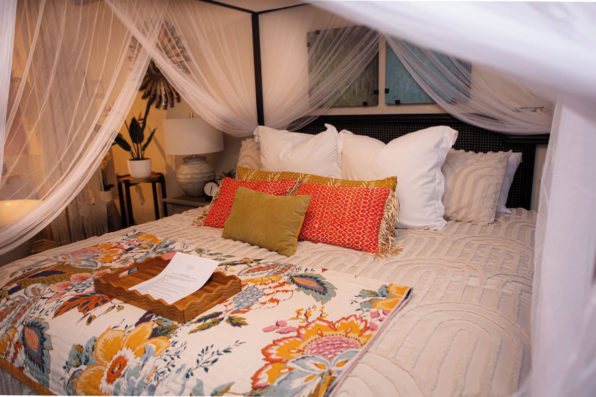 Una cama con dosel, una colcha estampada con motivos florales y una mesita de noche con lámpara.