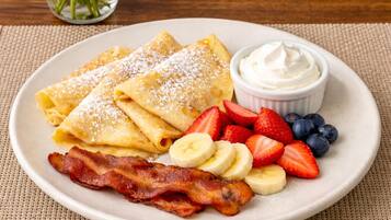 A plate of crepes with strawberries, blueberries, banana slices, and bacon, accompanied by a small bowl of whipped cream.