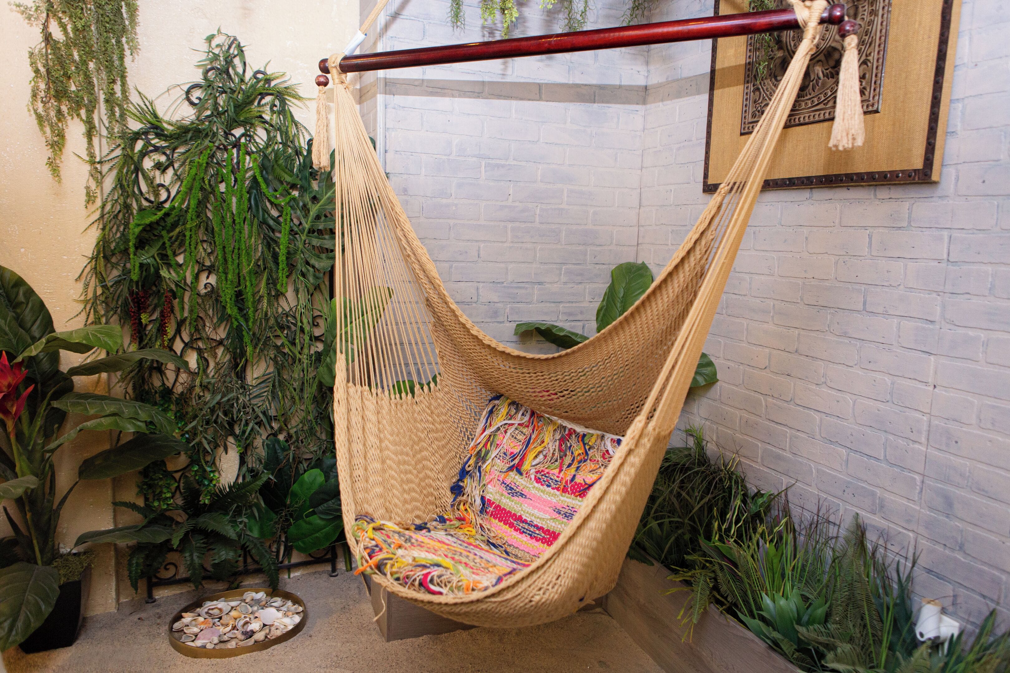 Un fauteuil suspendu en forme de hamac, agrémenté d’un coussin aux couleurs vives, entouré de différentes plantes et adossé à un mur de briques.