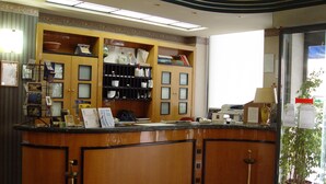 A reception desk with a wooden finish, a potted plant, and a large window providing natural light. - Hotel Kroma (Ragusa)